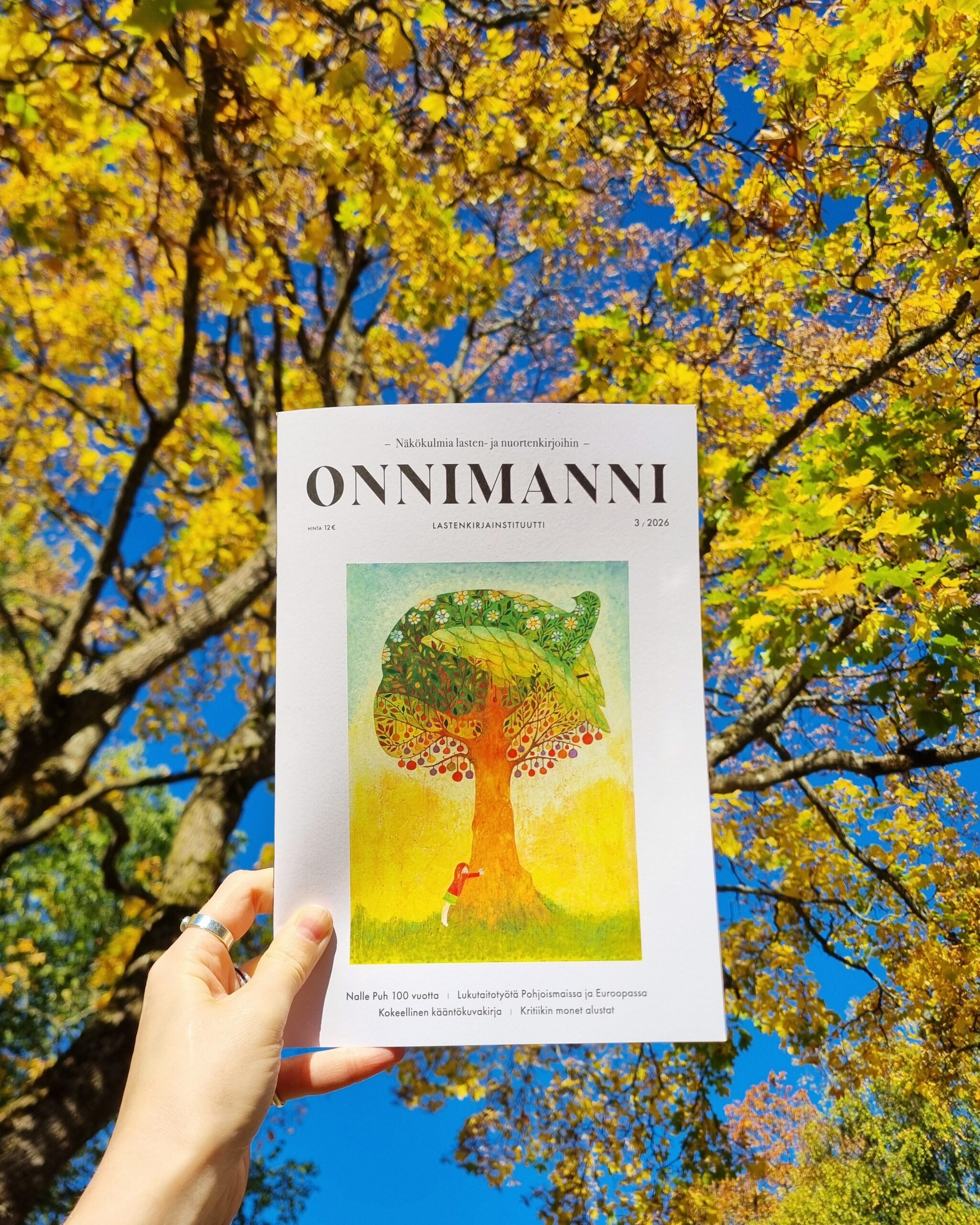 Käsi pitelee Onnimanni-lehteä, taustalla puunlatvat.